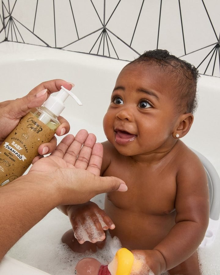 A parent pumps the baby shampoo while a smiling baby sits in a sudsy bath