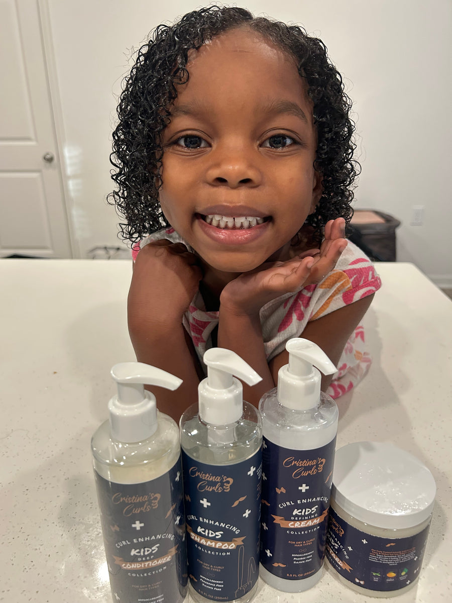 A baby sitting up on a bed, reaching for a jar from the Cristina's Curls routine