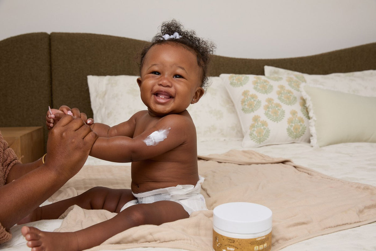 A laughing baby sitting on a bed with a smear of white cream on her upper arm, an adult hand holding her hand