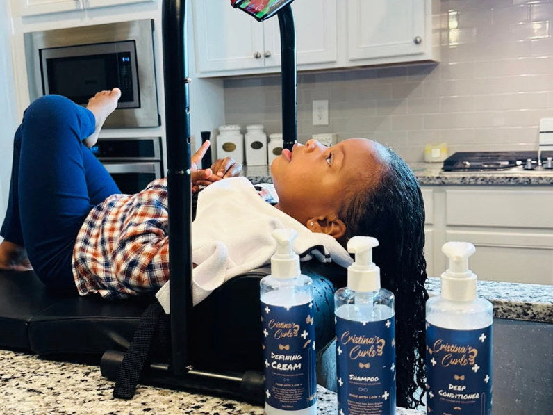 A child having her hair washed at a portable basin in a home kitchen, with three Cristina's Curls bottles in front