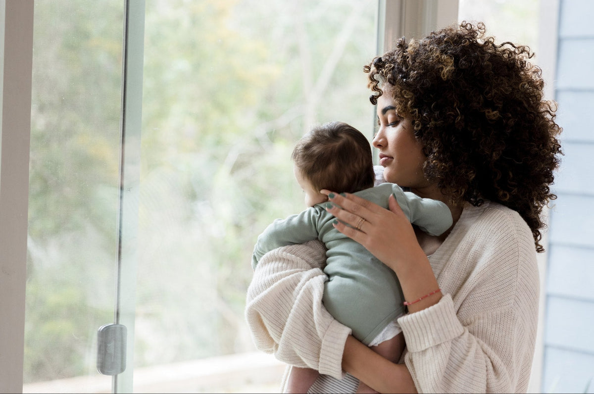 A mother holding her baby by a window, with the shampoo, conditioner and hair and scalp oil standing in front of them