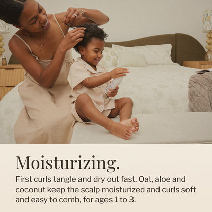 Mom styling a smiling toddler’s curls on a bed while the toddler holds the shampoo