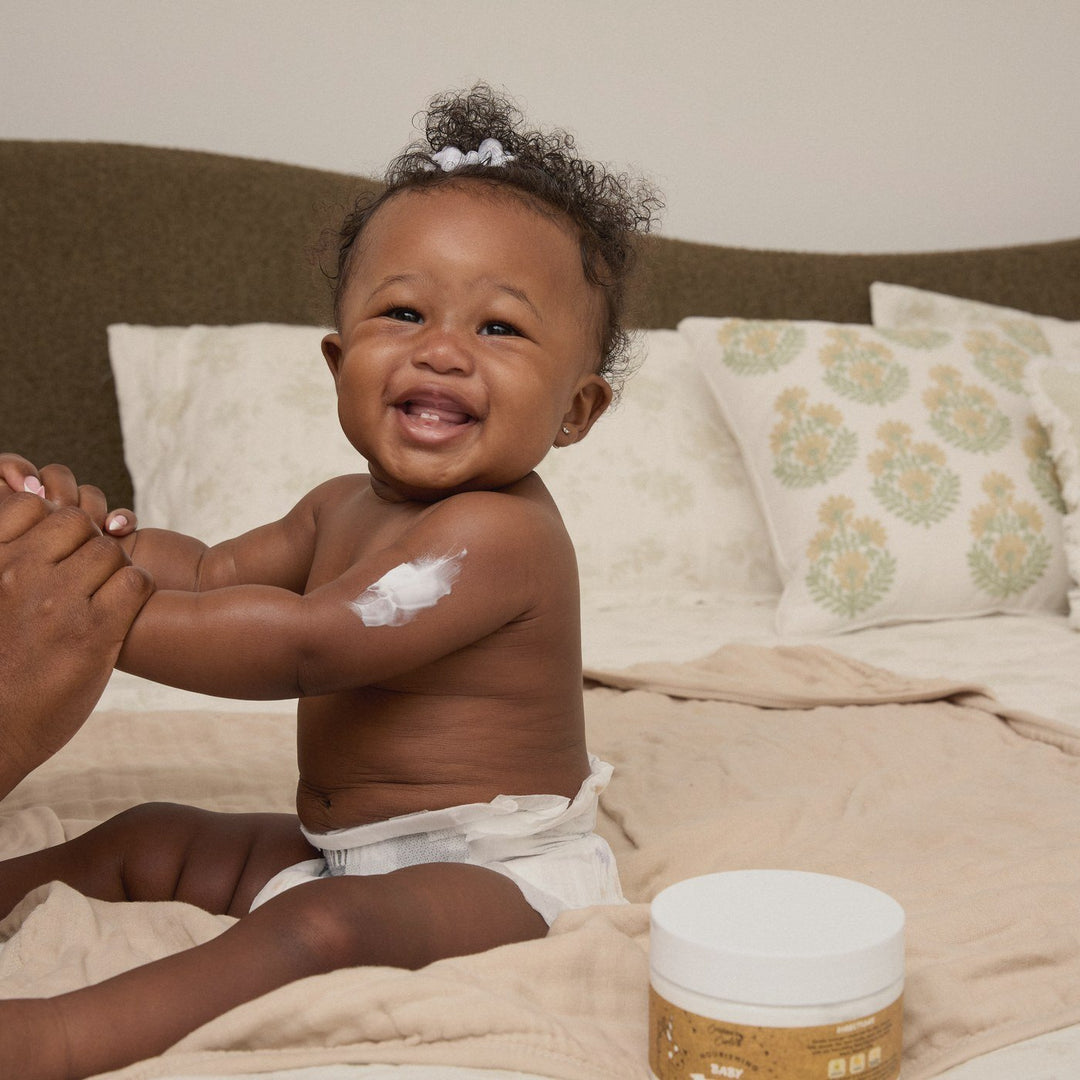 A smiling baby on a bed with a swipe of Body Cream on her arm, a parent's hands holding hers