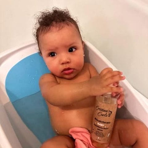 A baby in a bath seat holding the baby shampoo