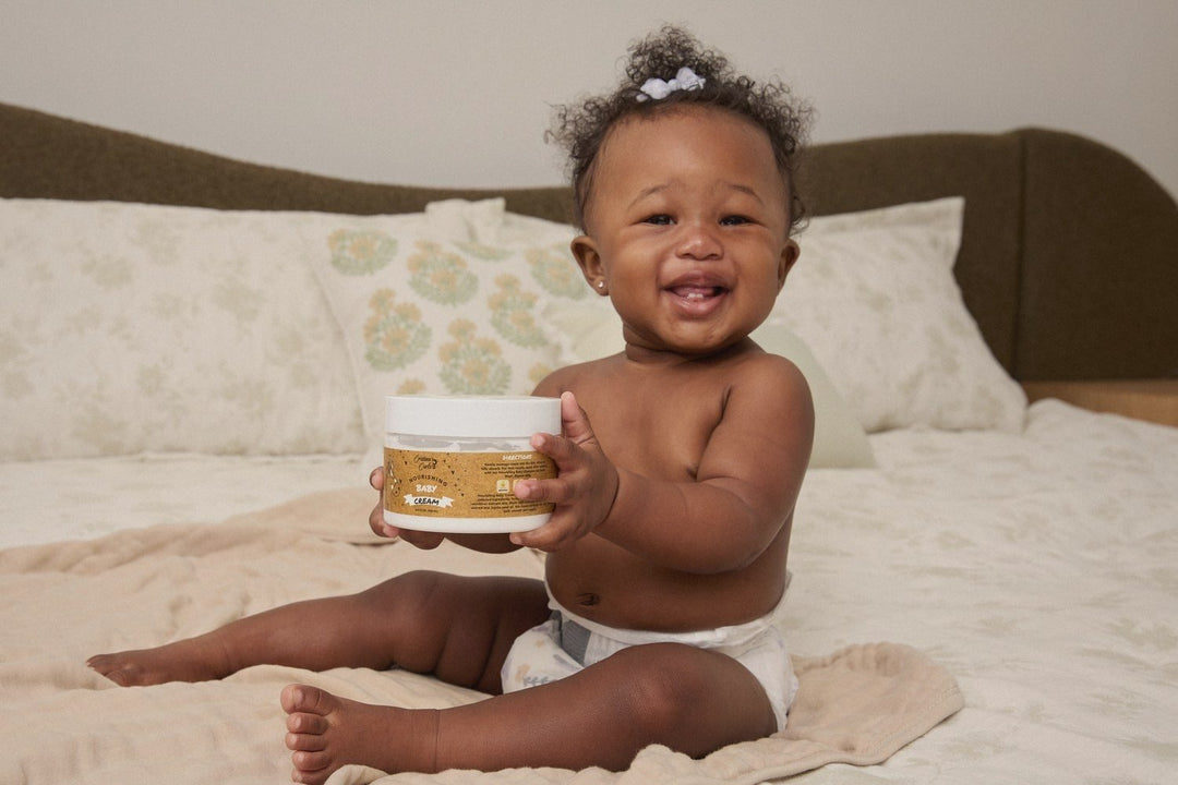A smiling baby on a bed holding the Body Cream jar